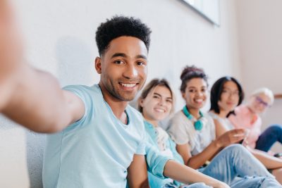 Handsome black young man with curly hairstyle making selfie with friends and smiling. Indoor portrait of joyful laughing students having fun after lesson and taking photo.