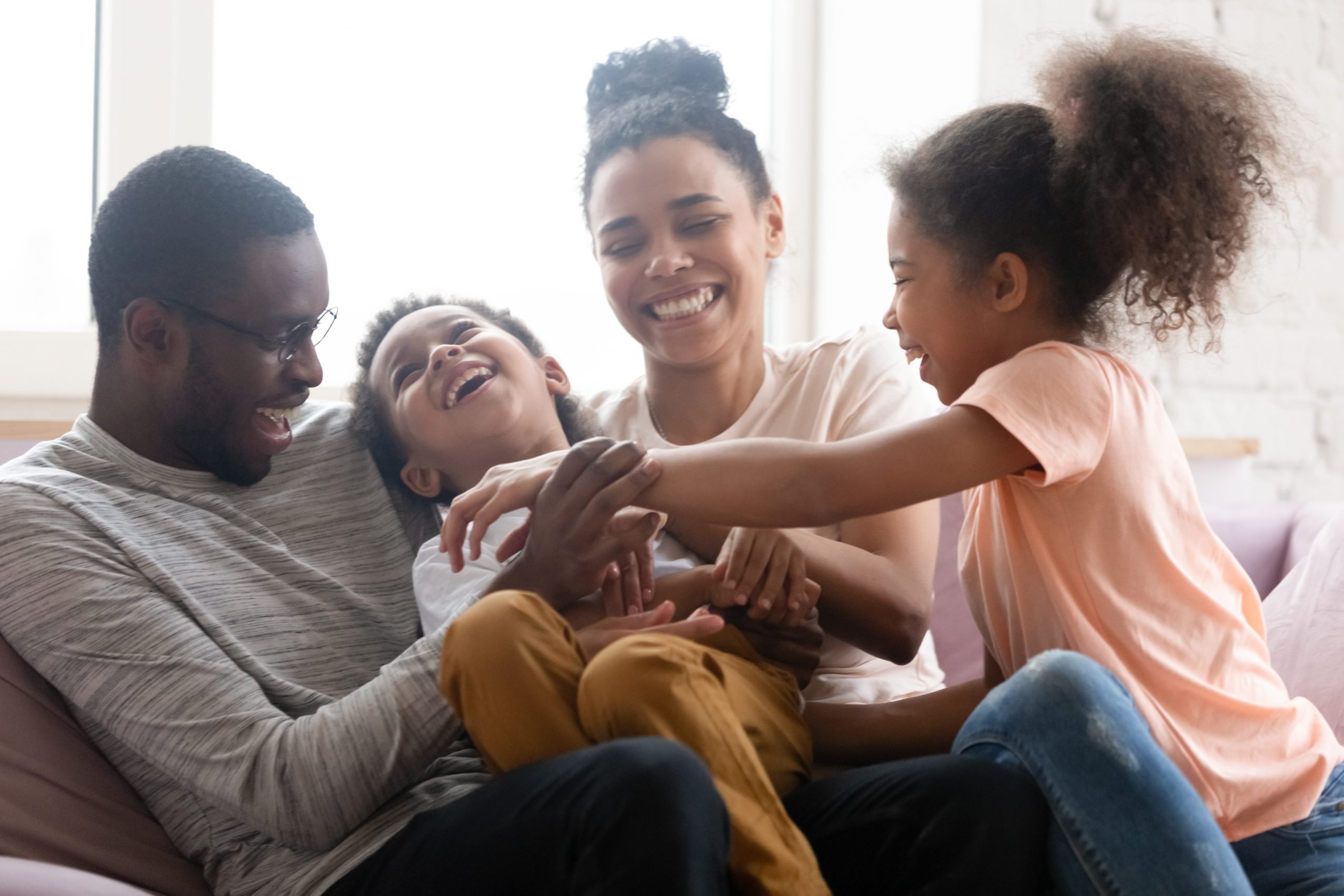 Overjoyed african American young family with little kids have fun playing relaxing on sofa in living room, happy biracial parents entertain with small children tickle laugh, enjoy weekend together