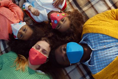 Top view of happy diverse family in safety mask relaxing on blanket during picnic. Mixed race parents and little children wearing protective mask lying on blanket outdoors