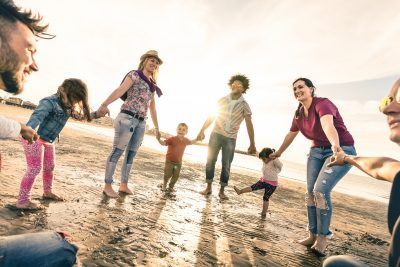 Happy multiracial families round dancing at the beach on ring around the rosy style - Multicultural happiness joy concept with mixed race people having fun outdoor at sunset