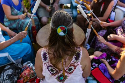 A high angle view of a woman wearing native headband and colorful clothes during a singing circle of people around a sacred mother drum outdoors.