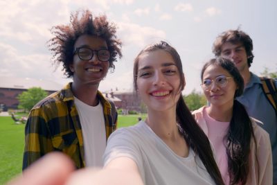 Pov shot of multiethnic young friends having video call or taking selfie together in park. Diverse young people looking at camera and talking standing outdoors