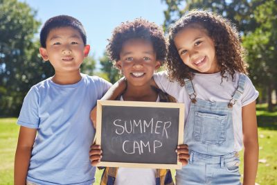Summer camp, portrait or children with board in park together for fun, bonding or playing in outdoors. Sign, diversity or happy young best friends smiling or embracing on school holidays outside.
