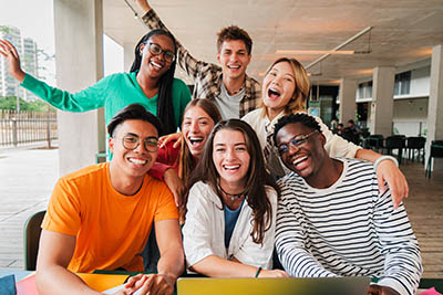 Group of young teenage students siting at library, using a laptop and looking at camera. Happy classmates studying and doing homework at high school. Multiracial academy people portrait. Schoolboys.