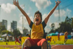 A smiling child sitting on a wheelchair celebrating while participating in a sporting event.