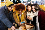 Young multiracial group of friends in winter clothes using mobile phone outdoor during christmas holidays.