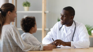 Smiling black male doctor listen cute african boy patient tell complaint make notes at medical consultation, mom and child son visit African pediatrician talk in clinic, children healthcare concept