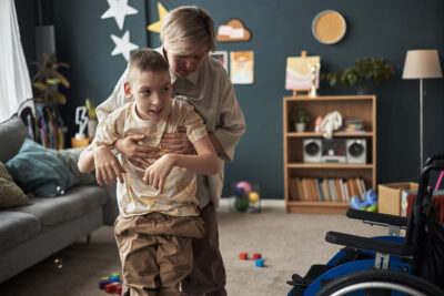 Caregiver assisting child with disability for physical exercise in a cozy living room filled with bookshelves, toys, and warm lighting emanating warmth and care