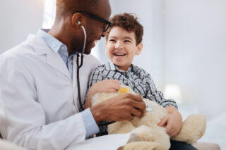 Immediate treatment. smiling male doctor sitting with boy while listening to plush bear's heart and talking.
