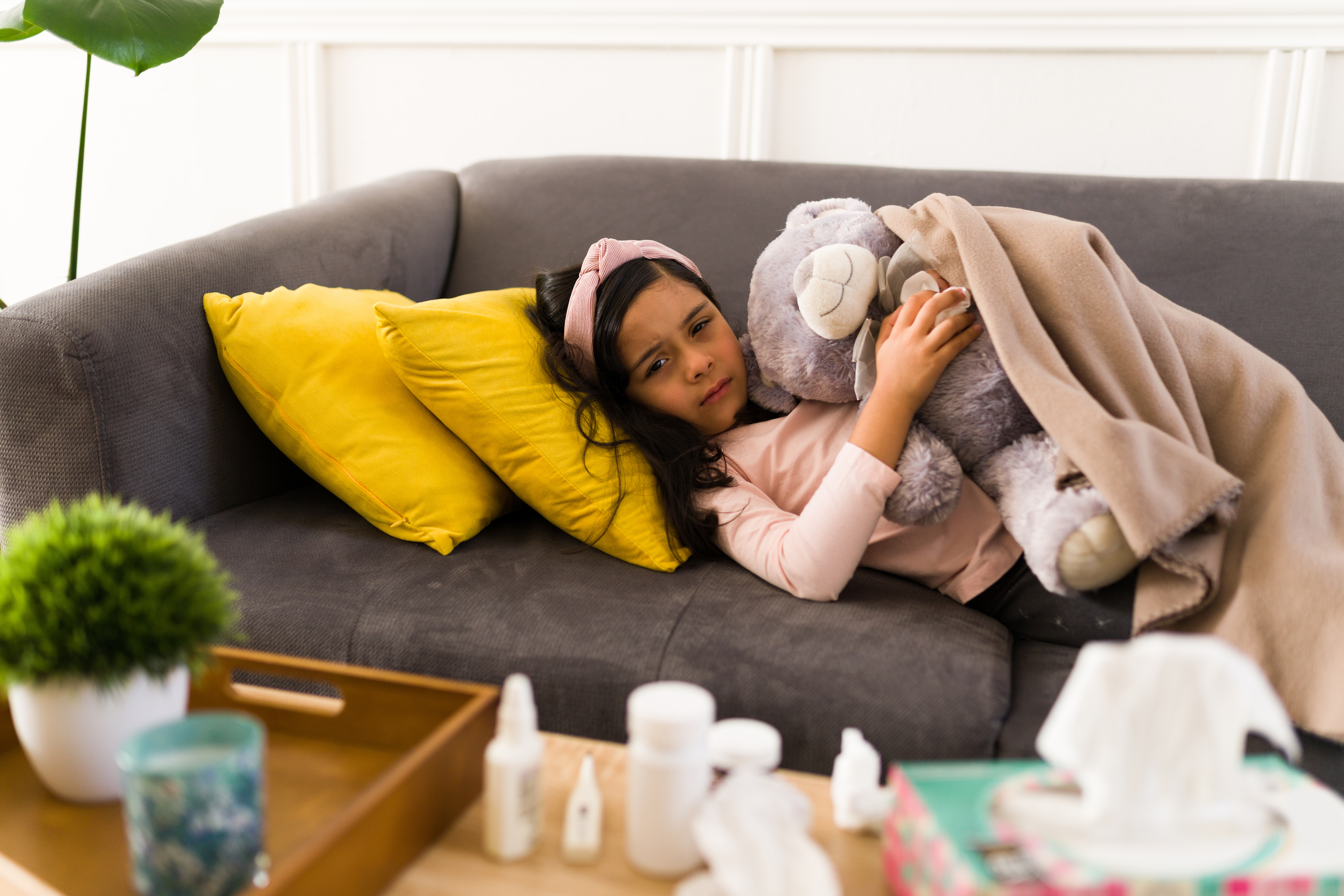 Portrait of a sad little girl hugging a teddy bear and covered with a blanket while recovering from a bad cold