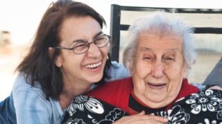 middle-age woman hugs elderly woman sitting in a chair. Both are smiling.