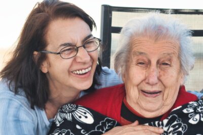 middle-age woman hugs elderly woman sitting in a chair. Both are smiling.