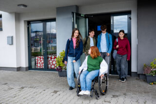 Group of teenage students supporting friend in wheelchair outside school building.