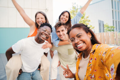 A lovely hispanic girl taking a selfie portrait with her multiracial teenage friends on piggybacks with cheerful attitude. Group of young student people laughing and having fun together outside